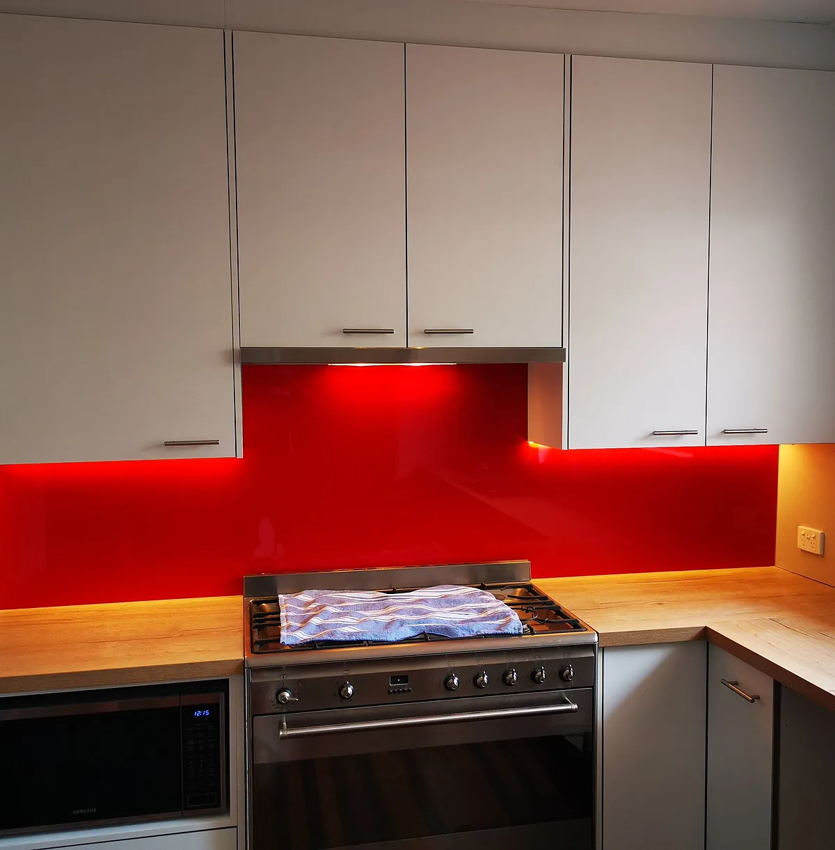Kitchen with red glass splashback lit by under-cabinet lighting above a freestanding oven